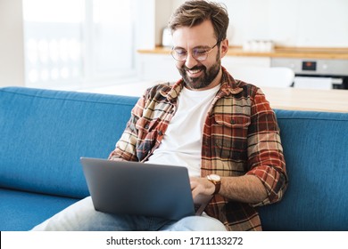 Portrait of young happy bearded man smiling and using laptop while sitting on sofa at home