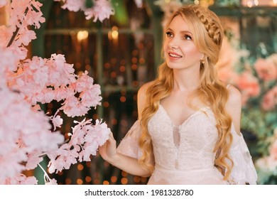 Portrait of young happy beautiful blonde woman in white vintage dress. Girl laughs, smile on face. Studio background, blooming pink sakura tree. Long hair is braided into an elegant romantic hairstyle