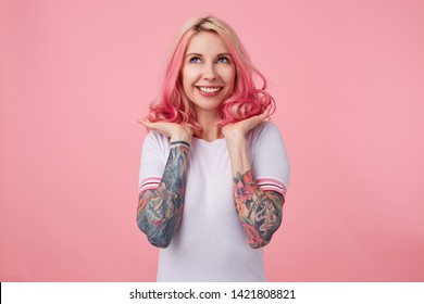 Portrait Of Young Happy Beautiful Pink Haired Lady With Tattooed Hands, Wears In White T-shirt, Straightens Her Hair, Happy With Her New Hair Color And Styling. Looks Up, Stands Over Pink Background.