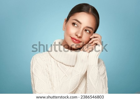 Portrait of young happy beautiful woman smiling and looking away, isolated on blue background in a warm white sweater. Concept of winter promo sale