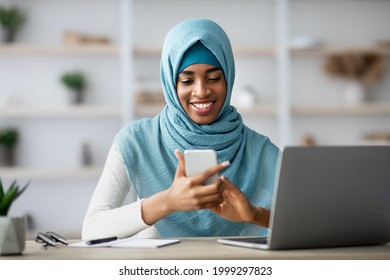 Portrait Of Young Happy Black Islamic Businesswoman In Hijab Using Cellphone While Sitting At Desk With Laptop, African Muslim Lady Texting On Mobile Phone While Working On Computer In Office