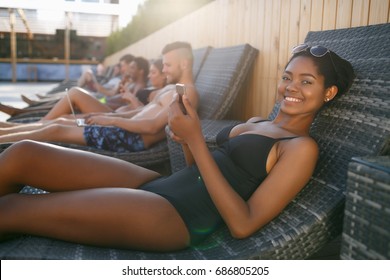 Portrait of young happy black sexy girl in bikini on deckchair with  smart phone near the pool next to her mixed race friends while traveling