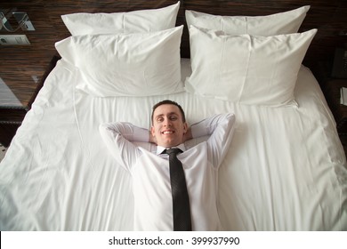 Portrait Of Young Happy Business Man Wearing White Shirt And Necktie Lying On The Bed In The Hotel Room With His Hands Behind Head. View From Above