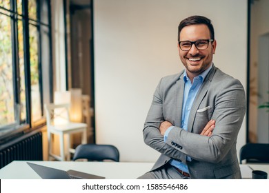 Porträt eines jungen, glücklichen Geschäftsmanns mit grauem Anzug und blauem Hemd, der in seinem Büro steht und mit gekreuzten Armen lächelt