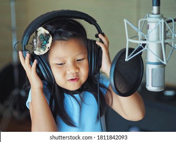 Portrait Of Young Happy Child Girl Singing Song With Headphone In Professional Voice Recording Studio With Microphone. Girl Singer And Kid Activities Concept.