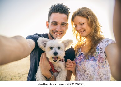 Portrait Of Young Happy Couple With Dog Taking A Selfie - Lovers On A Romantic Date On The Beach At Sunset