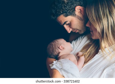 Portrait of young happy couple embracing and looking to their newborn over a dark background. Family and baby care concept.