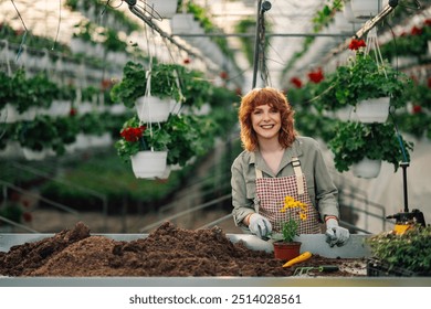 Portrait of young happy female ginger botanist standing at greenhouse with trowel in hands and planting blooming flower into a flowerpot while smiling at camera.Florist cultivating flowers at hothouse - Powered by Shutterstock