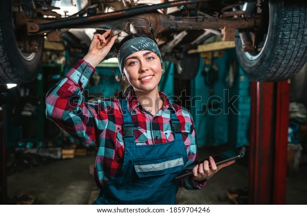 Portrait of a young happy female mechanic in\
uniform posing with tablet at her hands and remove glasses from the\
head. The car is located on the\
lift