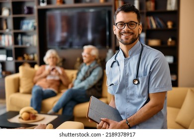 Portrait Of Young Happy Home Caregiver At Work Looking At Camera. Senior Couple In In The Background.