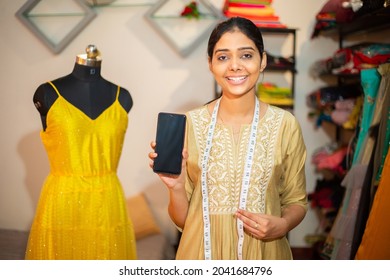 Portrait Of Young Happy Indian Woman Tailor, Female Smile Fashion Designer Showing Smartphone Blank Screen Or Display Mockup, Measurement Tap Around Neck In Garment Workshop Or Studio. Skill India, 