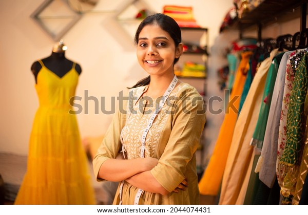 Portrait of Young happy indian woman, fashion\
designer tailor standing with crossed arms, measurement tape in\
garment workshop, seamstress, tailoring clothing on mannequin in \
studio. skill india