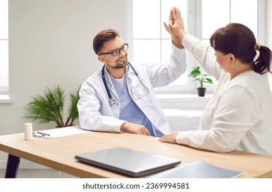 Portrait of a young happy male friendly doctor giving high five to a fat patient rejoicing success and reaching great results in treatment sitting at the desk in office examination room in clinic. - Powered by Shutterstock