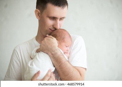 Portrait Of Young Happy Man Holding His Newborn Sweet Baby Dressed In White Clothes. Young Father Embracing His Baby With Love And Care, Smiling. Indoors Shot