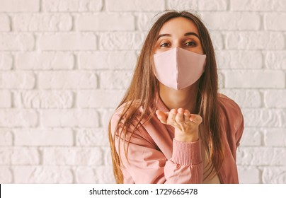 Portrait young happy millennial woman in trendy medical face mask blowing air kiss with hand. Beautiful stylish hipster girl in pink protective mask send blow kiss. Girl gesture blow kiss in face mask