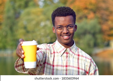 Portrait Of Young Happy Positive Man In Shirt And Glasses Holding Out A Plastic Cup Of Hot Drink Tea Or Coffee, Smiling, Looking At Camera. Black African Afro American Guy In Golden Autumn Park. 