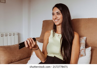 Portrait Of Young Happy Smiling Attractive Woman Watching Tv, Holds TV Remote Sitting At Couch At Home. Entertainment, Funny Series, Comedy Show Concept.