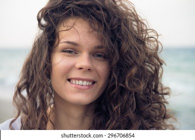 Portrait of young happy smiling woman on the coast in the summer. A woman with brown curly hair resting day on the beach in windy weather. Close up picture.