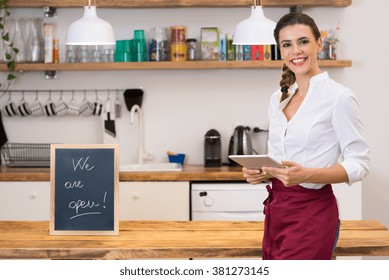 Portrait of young happy waitress looking at camera at bar with digital tablet. Portrait of a young female chef with blackboard in kitchen corner. Young smiling woman in her small business shop. - Powered by Shutterstock