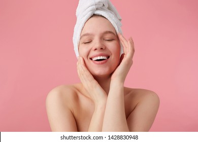 Portrait of young happy woman after shower with a towel on her head, broadly smiles with closed eyes, touches his face and smooth skin, stands over pink background - Powered by Shutterstock