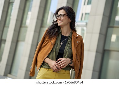 Portrait of a young happy woman holding smartphone, looking aside and smiling while walking on the city street on autumn day, stylish lady standing against blurred urban background