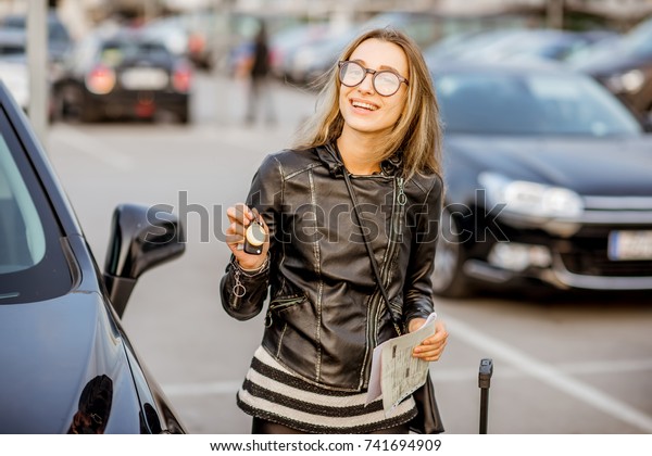 Portrait of a young happy\
woman with keys and rental contract standing near the car on the\
outdoor parking