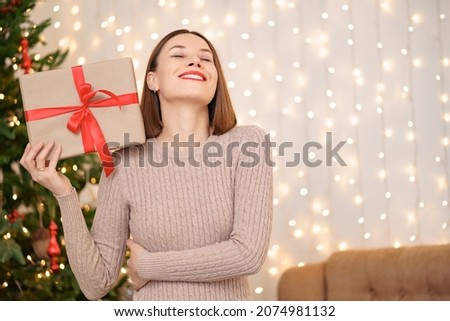 Portrait of young happy woman red lips looking at camera holding a wrapped gift box. Close up satisfied woman received present box. Festive Christmas lights background.