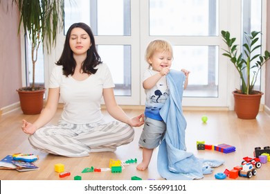 Portrait Of Young Happy Yoga Mom Spending Time With Her Little Baby Boy, Meditating In Lotus Position While Her Kid Son Playing Games At Home.