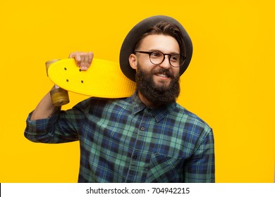 Portrait of young hipster man in hat and glasses holding yellow longboard and looking smilingly away. 