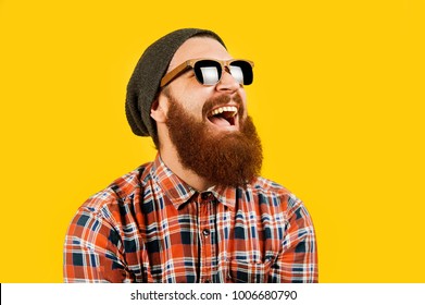 Portrait Of Young Hipster Man In Sunglasses And Hat Posing On Yellow Background. Smiling Bearded Man Wearing Sunglasses, Studio Shot. Happy Man With Beard Looking Up.