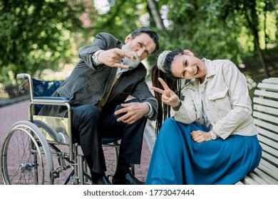 Portrait of young hipster modern girl with dreadlocks, sitting on bench, with the senior handicapped man in wheelchair, posing to camera in park, gesturing hand signs and having fun