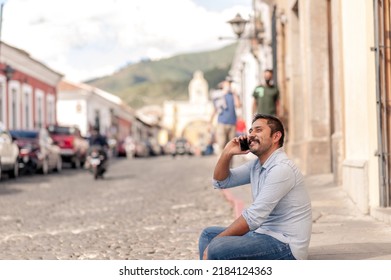 Portrait Of A Young Hispanic Man Enjoying His Vacations In A Colonial City. Hispanic Young Man Sitting On The Street Talking On The Cell Phone.