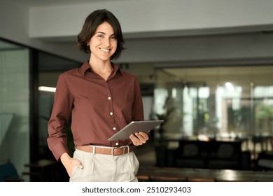 Portrait of young Hispanic professional business woman standing in office. Happy female company executive, smiling businesswoman entrepreneur corporate leader manager looking at camera using tablet.