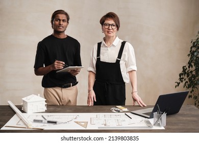 Portrait of young Indian architect with tablet and female engineer in bodysuit standing at table with supplies and blueprint