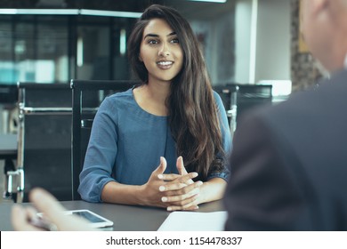 Portrait of young Indian female client or candidate sitting at table, talking to senior male manager and smiling in office. Job interview or consultancy concept