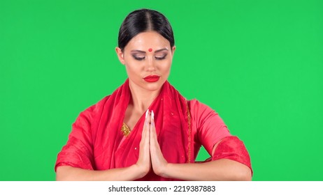 Portrait Of Young Indian Girl With Red Bindi Dot In National Classic Red Sari Looking Down. Brunette With Long Hair And Red Lips Folded Hands In Namaste Pose, Palm To Palm On Green Screen.