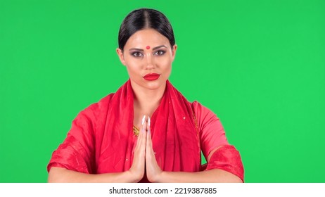 Portrait Of Young Indian Girl With Red Bindi Dot In National Classic Red Sari Looking Straight. Brunette With Long Hair And Red Lips Folded Hands In Namaste Pose, Palm To Palm On Green Screen.