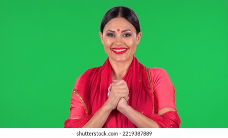 Portrait Of A Young Indian Girl With Red Bindi Dot In National Classic Red Sari Looking Straight, Smiles And Folded Palm To Palm. A Brunette With Long Hair And Red Lips On Green Screen At Studio.