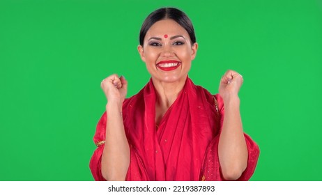 Portrait Of Young Indian Girl With Red Bindi Dot In National Classic Red Sari Looking Straight, Smiles And Folded Palm To Palm. Brunette With Red Bindi Dot In National Classic Red Sari On Green Screen