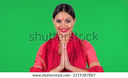 Portrait of a young Indian girl with red bindi dot in national classic red sari looking straight, smiles and folded palm to palm. A brunette with long hair and red lips on green screen at studio.