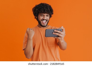 Portrait Of Young Indian Handsome Curly Enthusiastic Man Holding His Phone Celebrating Victory In Mobile Game With Raised Fist Over Isolated Orange Background