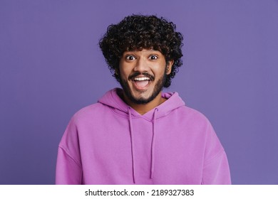 Portrait Of Young Indian Handsome Curly Enthusiastic Man Wearing Violet Hoodie Looking At Camera With Opened Mouth While Standing Over Isolated Violet Background