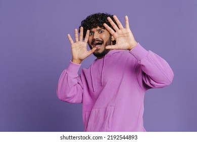 Portrait of young indian handsome curly scared man stretching arms showing surrender gesture , and looking at camera standing over isolated violet background