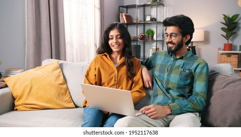 Portrait of young Indian happy cheerful family couple resting on sofa in living room searching internet typing online on laptop computer and speaking, choosing decor, e-commerce, leisure concept - Shutterstock ID 1952117638