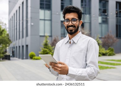 Portrait of a young Indian man in glasses and a white shirt standing near an office building, holding a tablet and smiling at the camera.