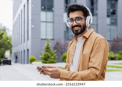 Portrait young Indian man with headphones and tablet watching online video sitting on bench near office building businessman smiling and looking at camera, portrait of satisfied online stream viewer - Powered by Shutterstock