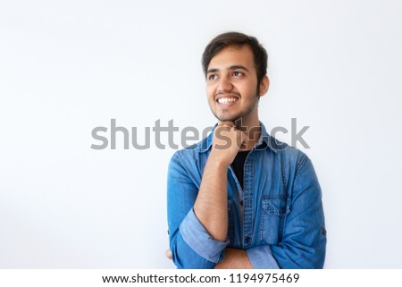 Portrait of young Indian man looking upwards and smiling. Positive handsome dark haired man wearing denim shirt propping up chin with right hand. Happy memory concept.