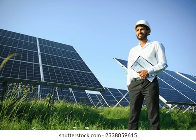 Portrait of Young indian man technician wearing white hard hat standing near solar panels against blue sky.Industrial worker solar system installation, renewable green energy generation concept - Powered by Shutterstock