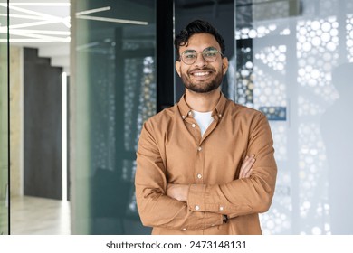 Portrait of a young Indian man wearing glasses and a brown shirt standing in a modern office, crossing his arms over his chest and looking confidently at the camera.