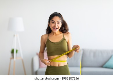 Portrait of young Indian woman measuring her waist with tape indoors, smiling at camera. Beautiful slim Asian lady having fit body with perfect parameters. Healthy diet and weight loss concept